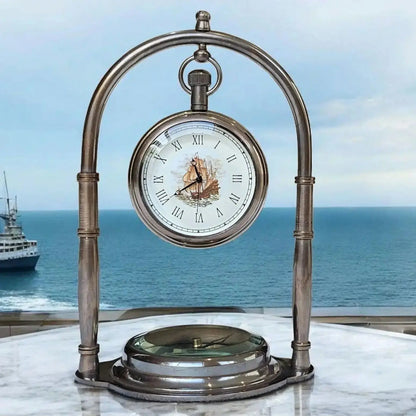 A vintage brass arch clock with a ship dial, featuring a white face with black Roman numerals, housed in a metal structure with a base, against a maritime-themed backdrop with a boat.